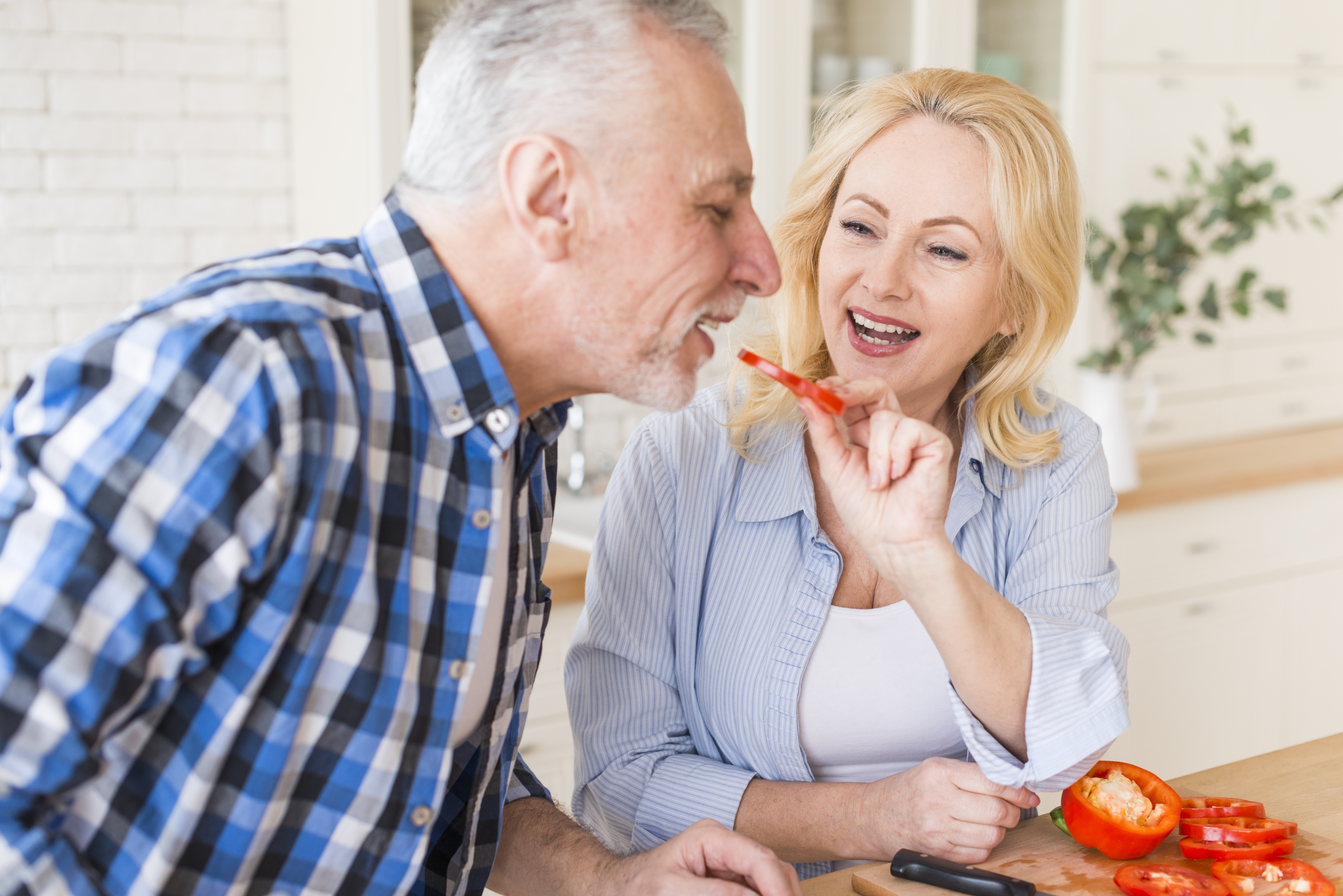 happy-portrait-senior-woman-feeding-slice-bell-pepper-her-husband-kitchen